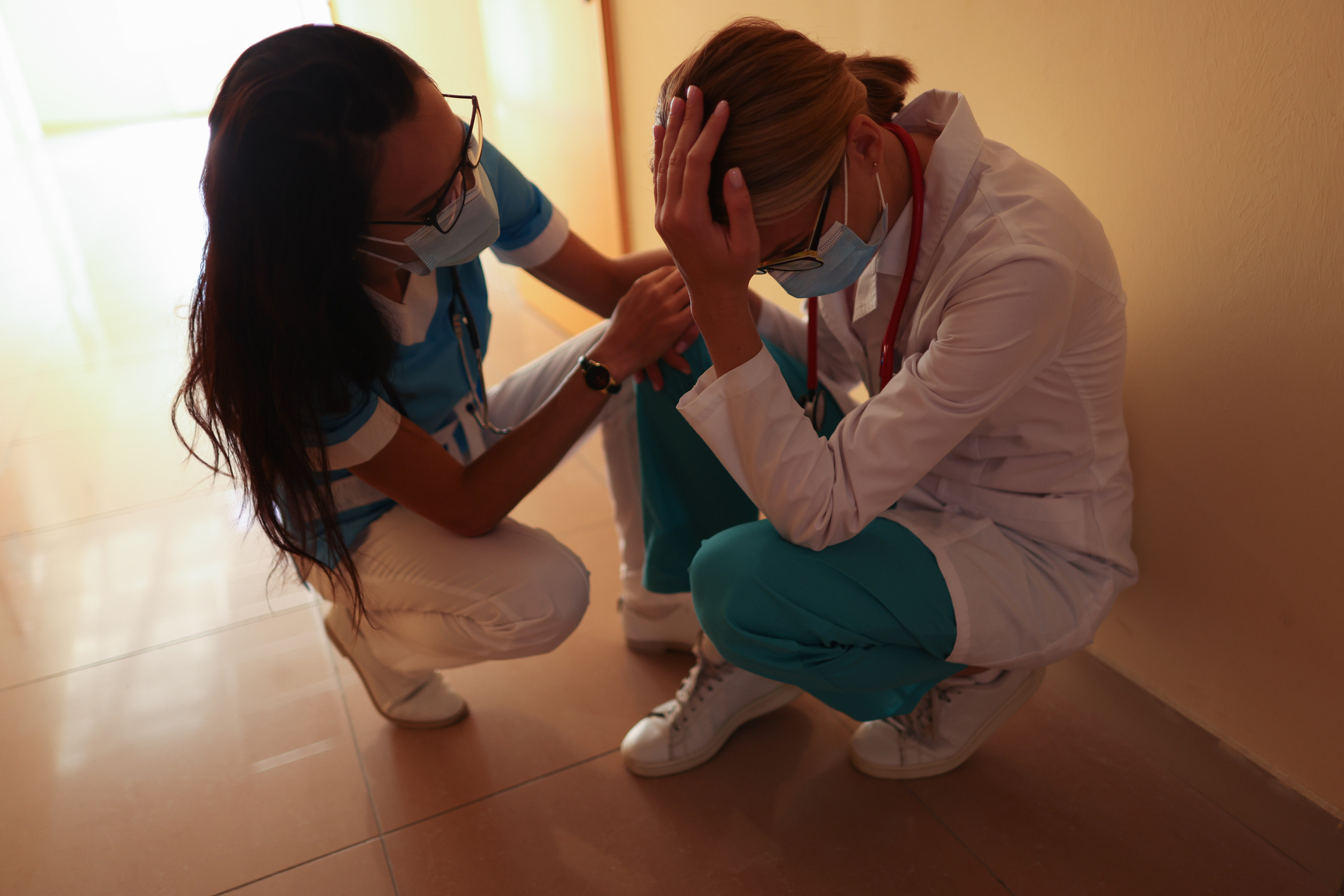 Nurse calms crying doctor wearing medical protective mask in bright hospital corridor.
